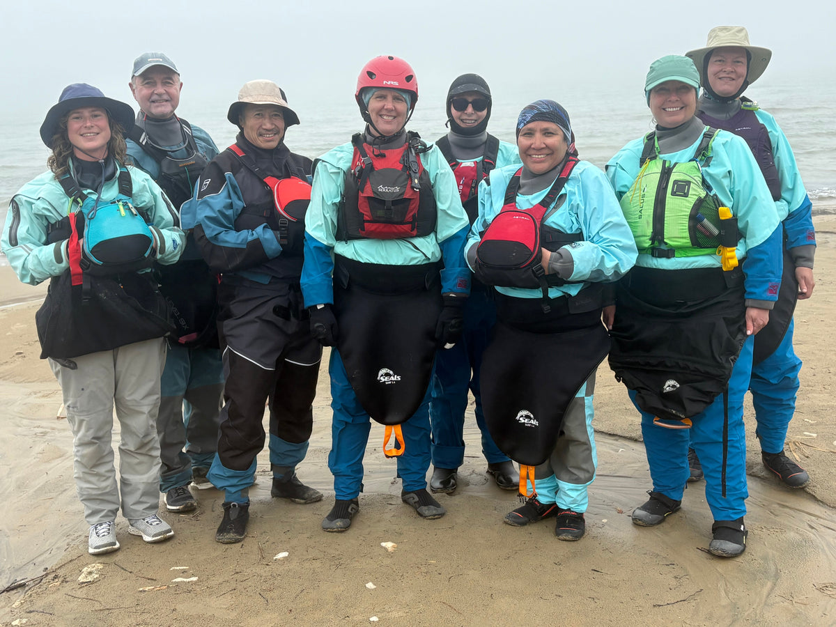 A group of smiling sea kayakers in drysuits and PFDs on a beach